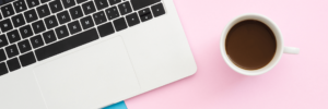 A macbook pro keyboard next to a full cup of coffee both resting on top of a pink background with some blue showing as well
